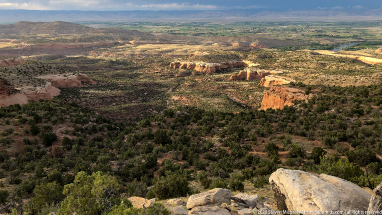 Pale foreground boulders overlook a scrub-covered basin and sunlit red sandstone cliffs.