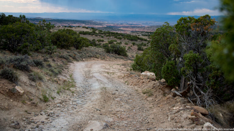 A rough dirt trail descends through juniper and sage toward distant mesas.