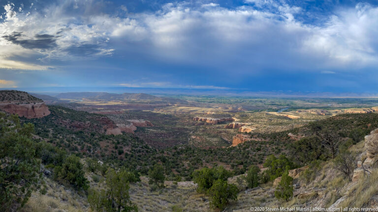 Dark clouds send rain onto a distant plateau above a scrub-covered canyon basin.