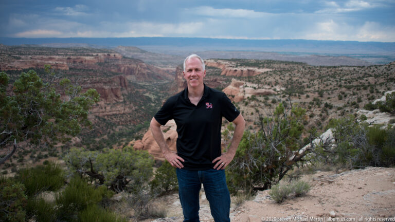 A man in a black polo stands among shrubs at a canyon overlook with red cliffs behind.