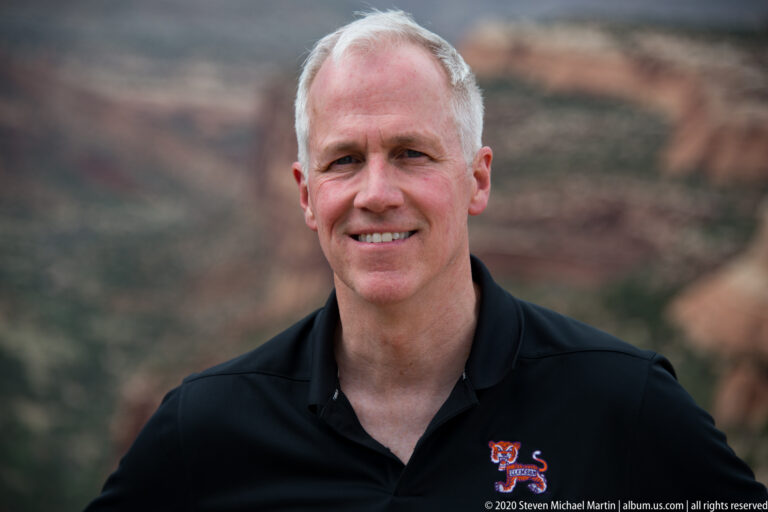 A smiling man in a black polo is shown in close portrait against a blurred canyon backdrop.