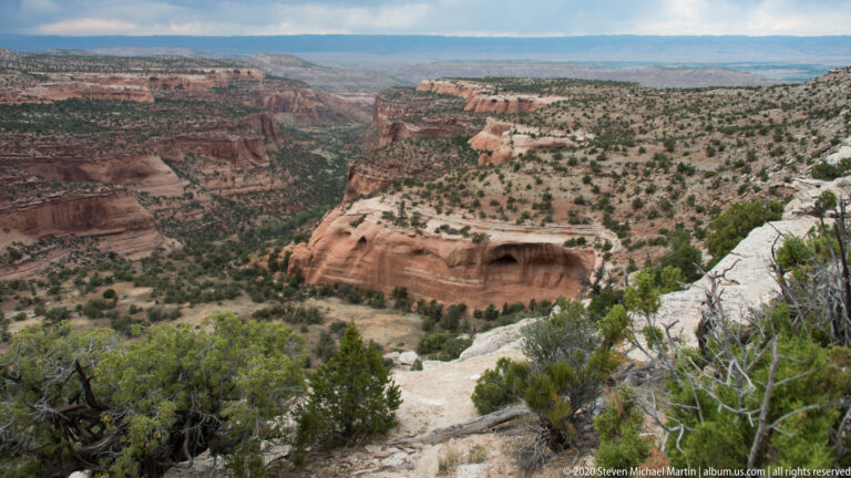 A dark alcove cuts into a red sandstone wall across a ravine, with slickrock in the foreground.