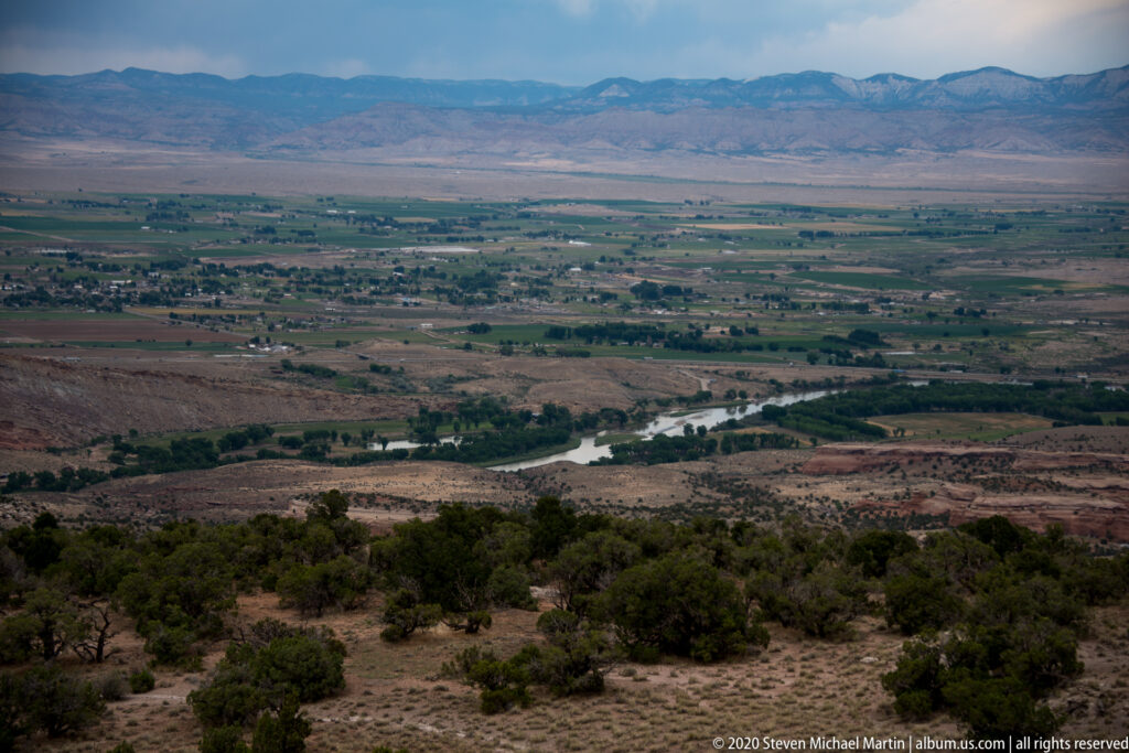 A river winds through green fields beneath distant mountains, viewed from a dry canyon rim.