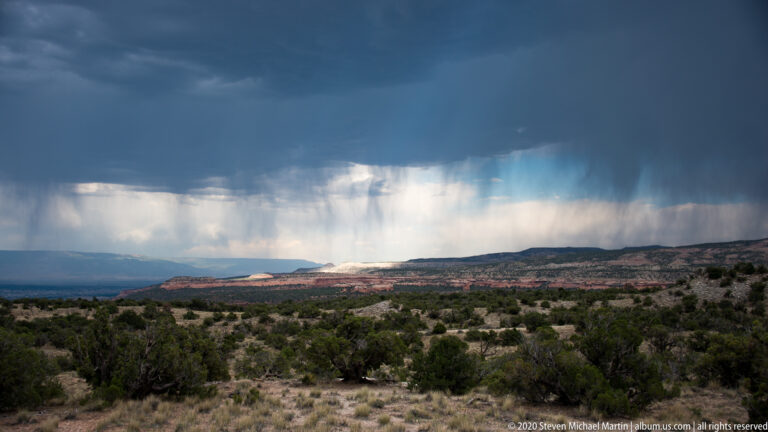A rain shaft falls onto distant plateau beneath dark clouds beyond dry foreground scrub.