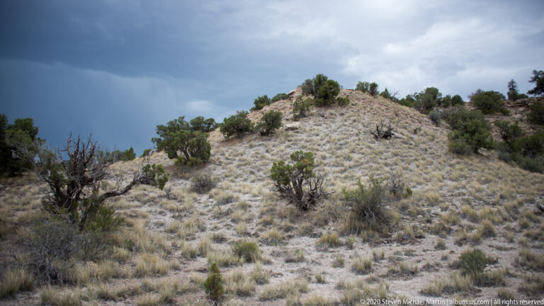 A pale shrub-covered hill rises beneath dark storm clouds with scattered junipers on its slope.