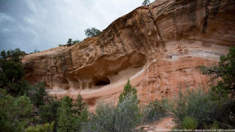 A dark shallow cavity lies beneath a curved, banded sandstone overhang above desert shrubs.