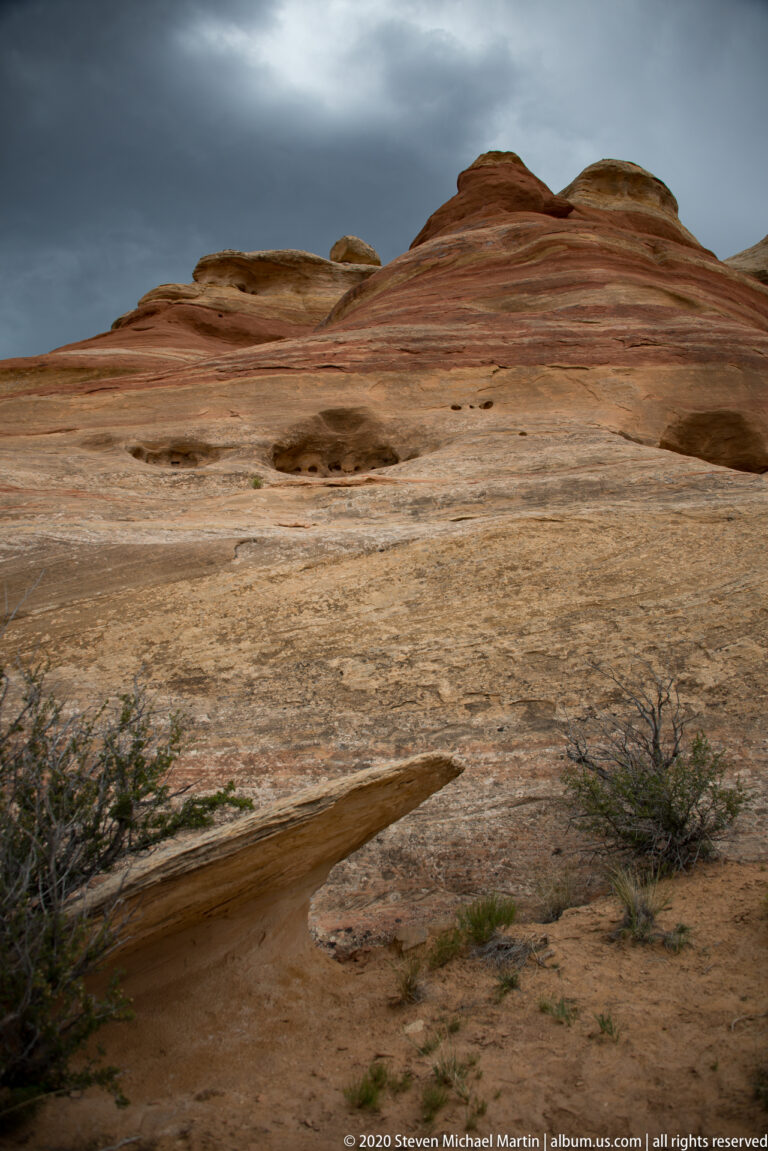 Horizontal red and tan bands cross a broad smooth sandstone bench beneath darkening clouds.