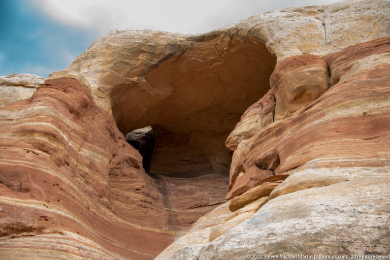A rounded dark alcove sits beneath a pale sandstone arch amid curved red strata.