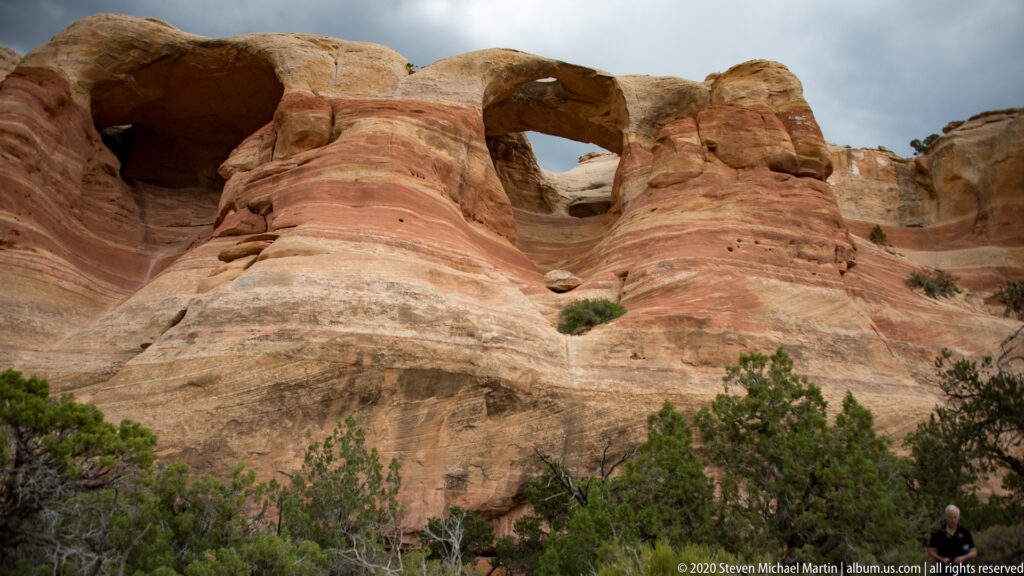 Two sandstone openings pierce a broad ridge above shrubs, with a visitor at the base.