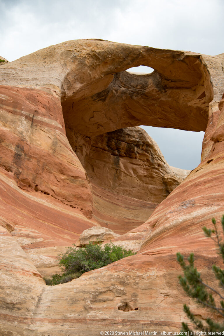 A broad sandstone arch spans a rounded rock basin with shrubs on the lower slope.