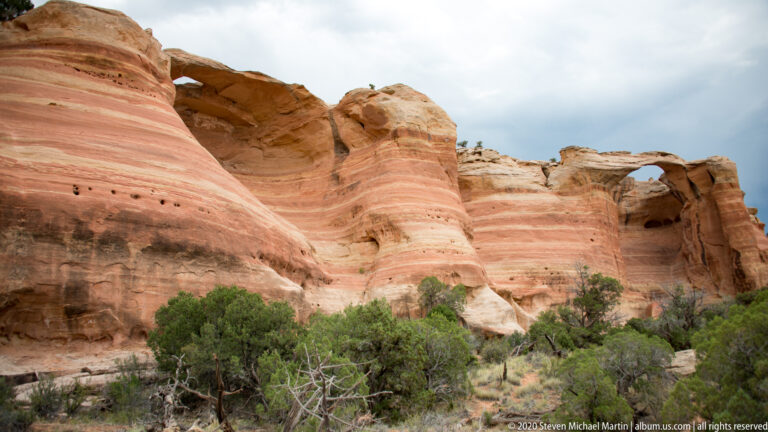 A dark alcove and smaller arch interrupt layered sandstone above desert shrubs.
