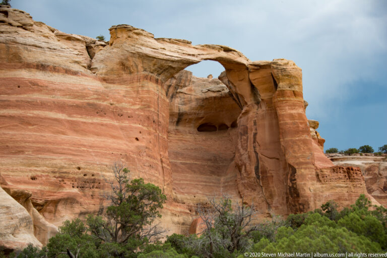 A high sandstone arch pierces a rounded buttress above brush and pale sand.