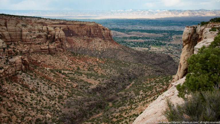A rocky canyon descends toward a greener developed valley between red and cream cliffs.