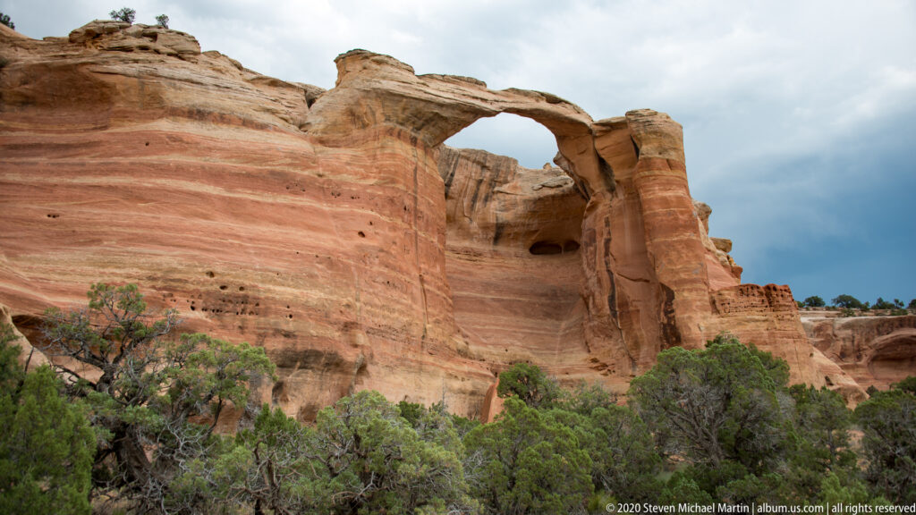 A visitor stands below a broad sandstone arch on a sandy path among shrubs.