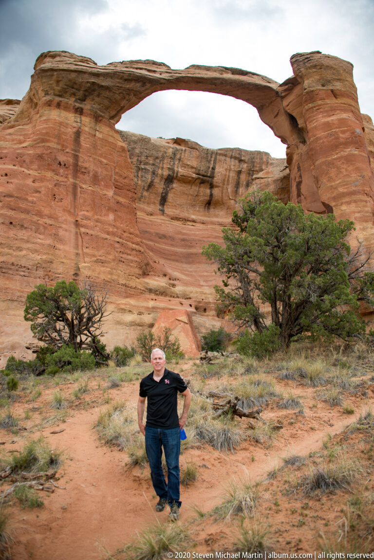 A visitor stands on a sandy path beneath a broad sandstone arch and red cliffs.