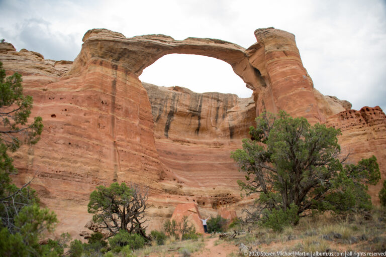 A natural sandstone arch spans layered red cliffs above shrubs and sandy terrain.