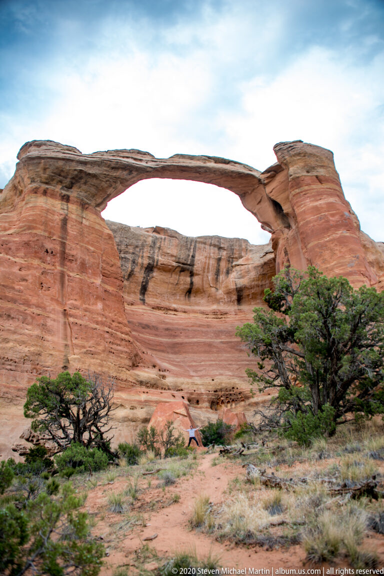 A high sandstone arch crosses a layered wall above a scrub-covered slope.