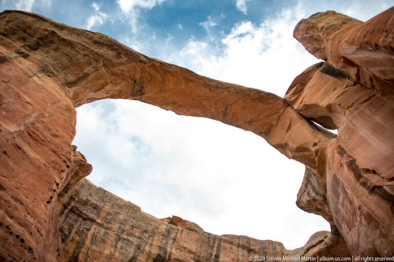 The underside of a sandstone arch sweeps overhead between red rock walls and clouds.