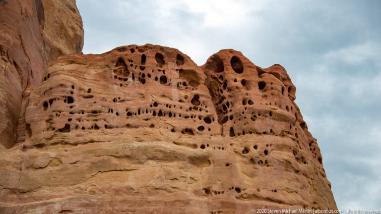 A sandstone tower covered in rounded cavities rises above a lower ledge beneath clouds.