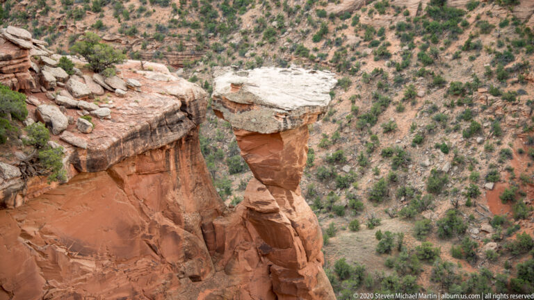 A pale capstone balances on a narrow sandstone pedestal above a canyon slope.