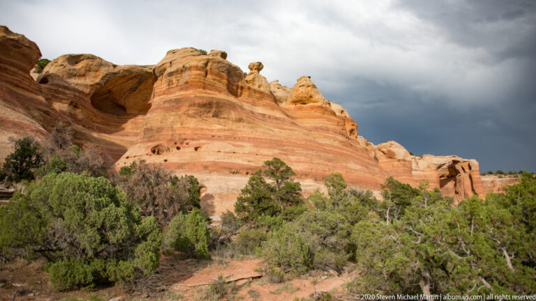 Rounded sandstone pinnacles rise behind junipers beneath a dark, cloudy sky over sandy ground.