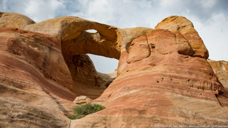 A curved sandstone arch spans two layered rock walls beneath cloudy sky.