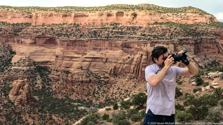 A visitor photographs layered red canyon walls from a rocky overlook above the valley.