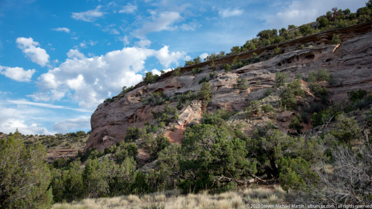 A layered sandstone cliff rises over shrubs and small trees beneath a blue sky with scattered clouds.
