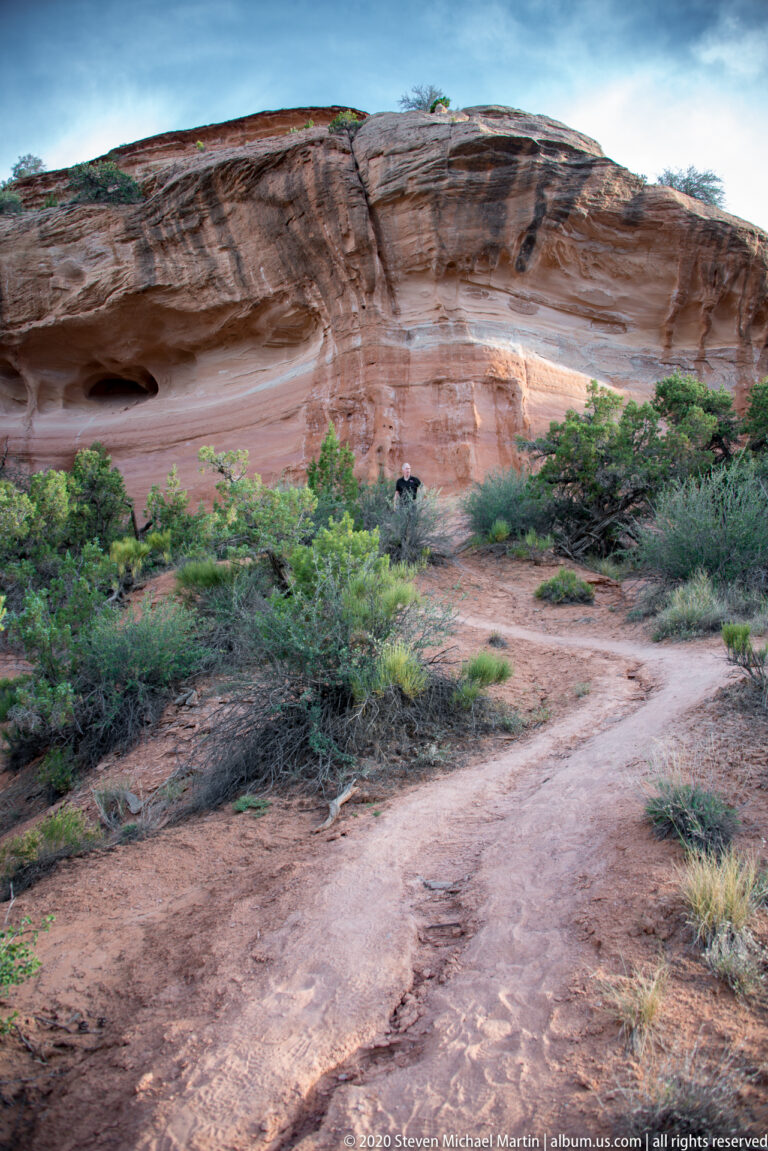 A sandy trail curves through low shrubs toward a broad sandstone wall with several dark openings.