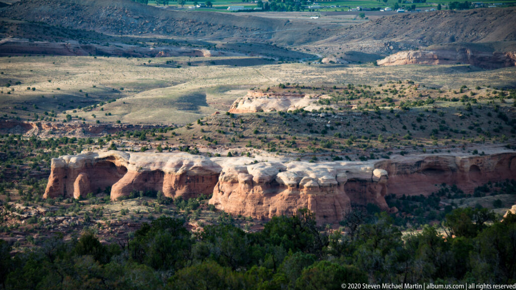 A pale sandstone cliff band with several dark openings crosses a broad, sparsely vegetated basin.
