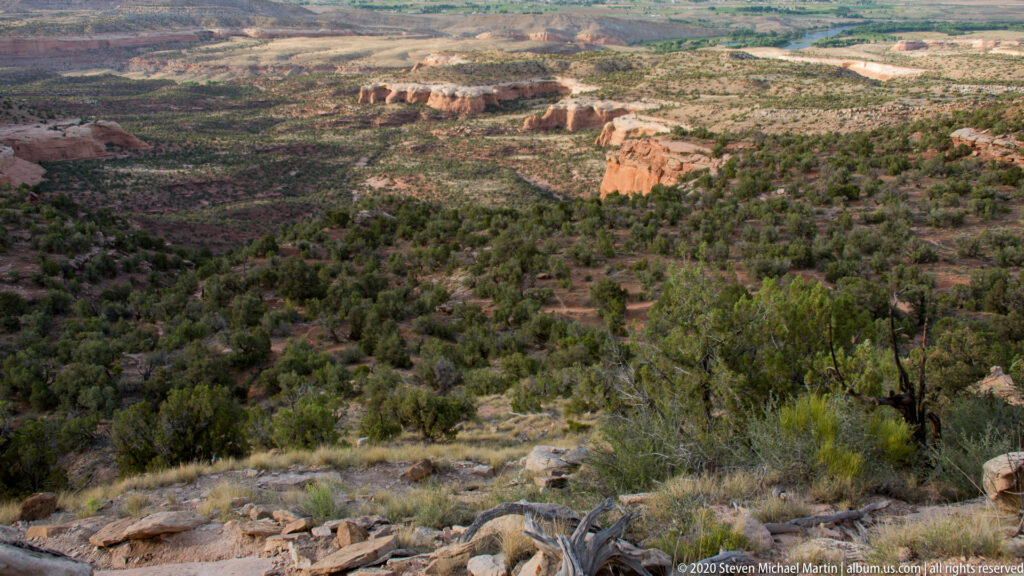 Red sandstone cliffs and isolated rock towers rise beyond a scrub-covered slope, with mesas fading into the distance.