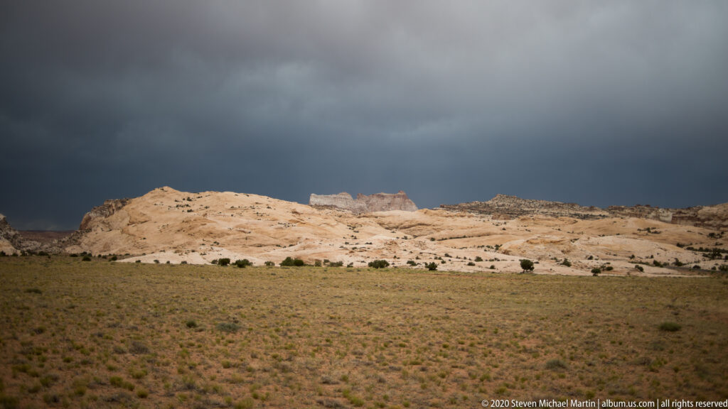 A pale rock escarpment stands beyond open scrub beneath a dark storm front.