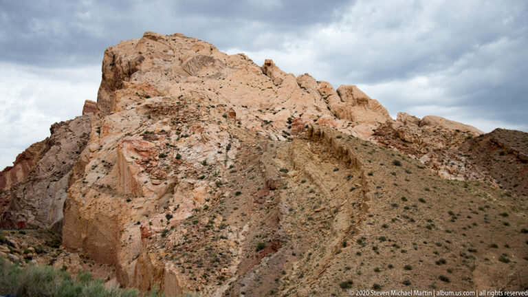A fractured orange sandstone cliff rises above sparse shrubs on a steep slope.