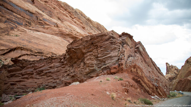 A rust-colored layered cliff rises over red talus beside a narrow road opening.