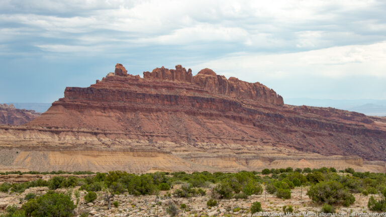 A red-capped ridge with small pinnacles rises above pale layered slopes and scrub.
