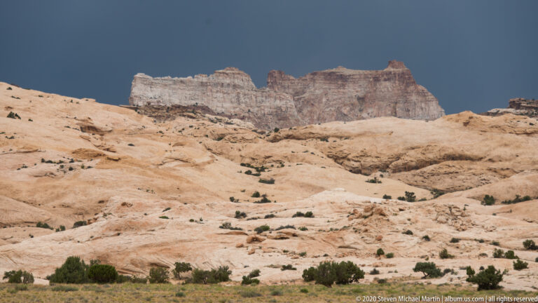 Pale sandstone benches and shallow channels slope below a darker horizontal cliff.