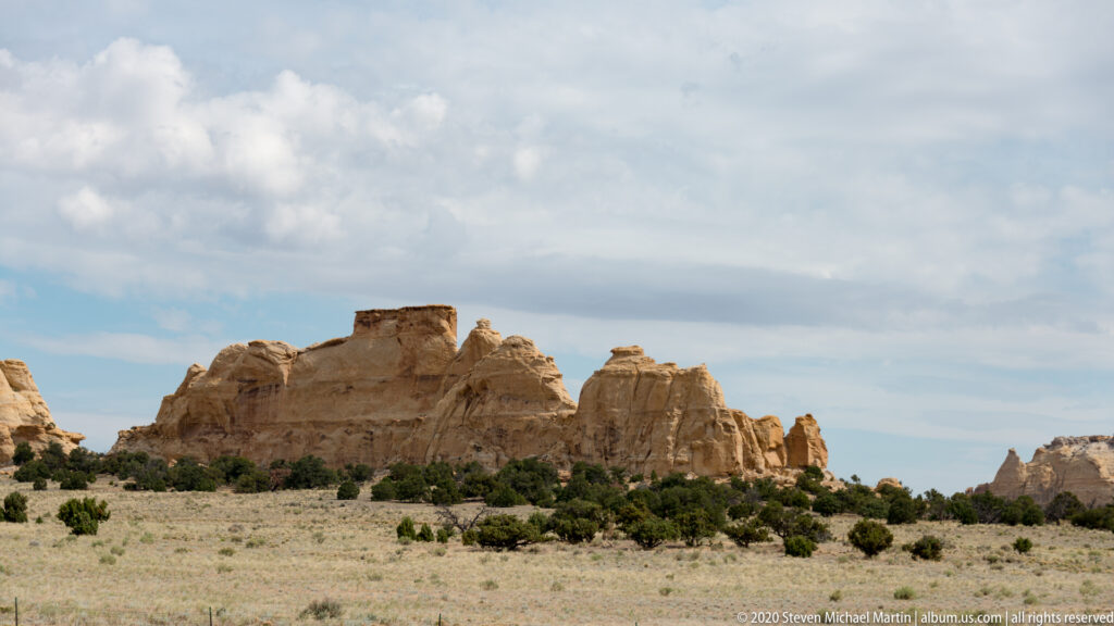 Pale rounded sandstone towers rise behind junipers above an open sandy plain.