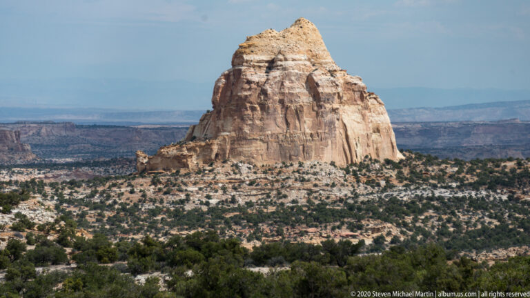 A pale rounded butte rises above juniper scrub with distant mesas behind it.