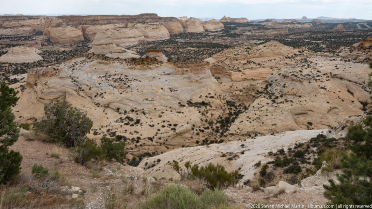 A pale rock bench descends into a shrub-dotted basin beneath layered cliffs.