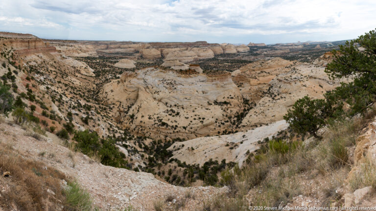 A wide pale canyon basin with scattered shrubs lies below layered cliffs.
