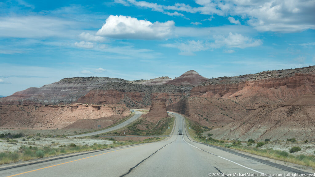 Two-lane highway winds between layered red and gray desert ridges beneath a partly cloudy sky.