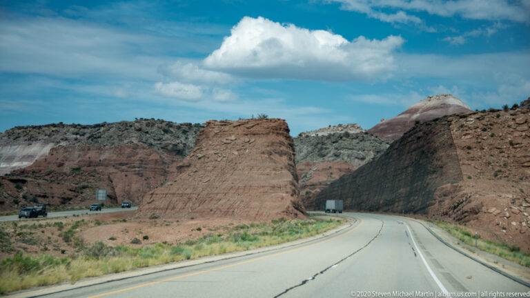 Highway curves around a flat-topped red butte with a pale rounded summit behind it.