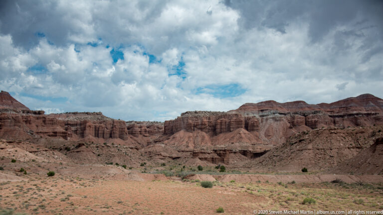 Open sandy basin with sparse shrubs sits below layered red and tan cliffs beneath large clouds.