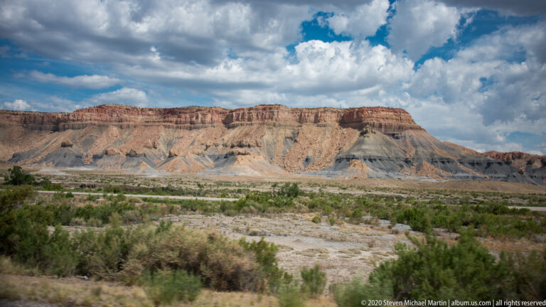 Layered red, tan, and gray cliffs rise above a scrub plain beneath large white clouds.