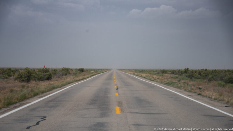 Straight two-lane highway with yellow center lines disappears into a hazy horizon between low shrubs.