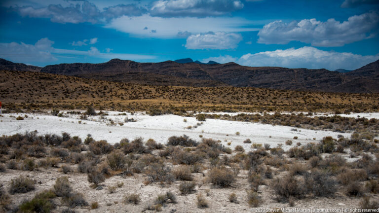 Pale flat basin lies beyond desert shrubs beneath a dark layered ridge and scattered clouds.