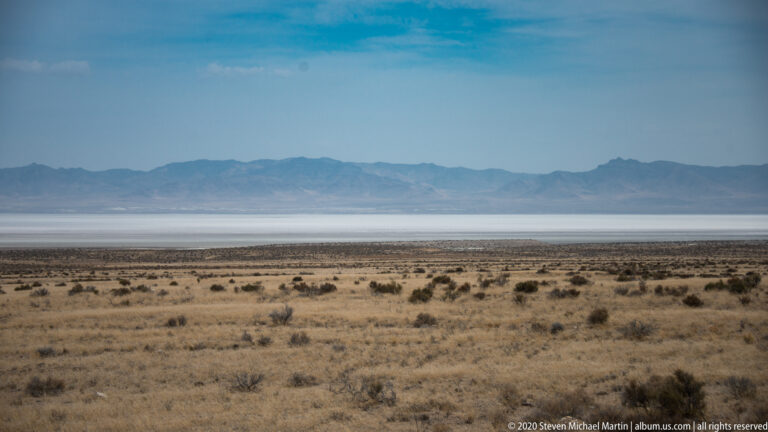 Sparse desert scrub extends toward a pale flat and distant blue mountains beneath a hazy sky.