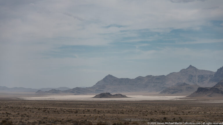 Jagged mountain peaks stand beyond a pale basin and sparse desert scrub in the foreground.