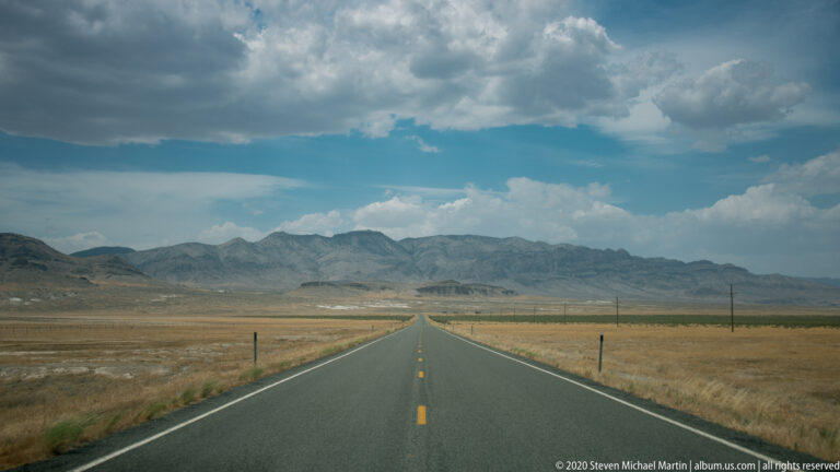 Paved highway crosses dry open ground toward distant mountains beneath large clouds.