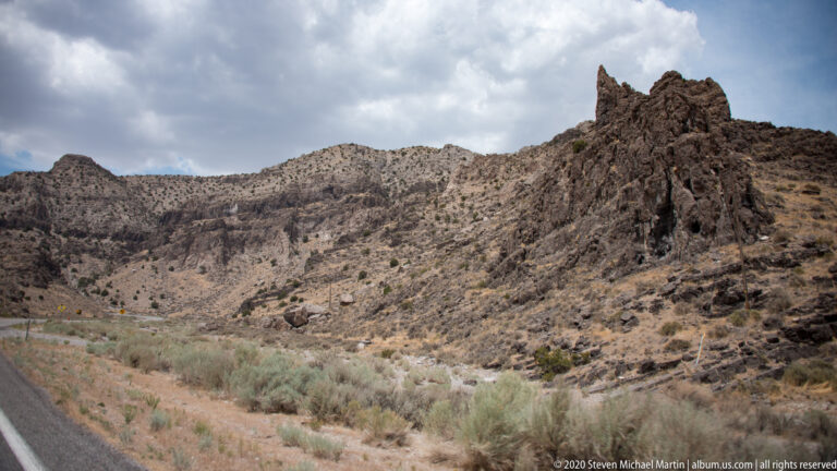 Jagged reddish-brown rock ridge rises above a pale shrub-covered slope beside a road.